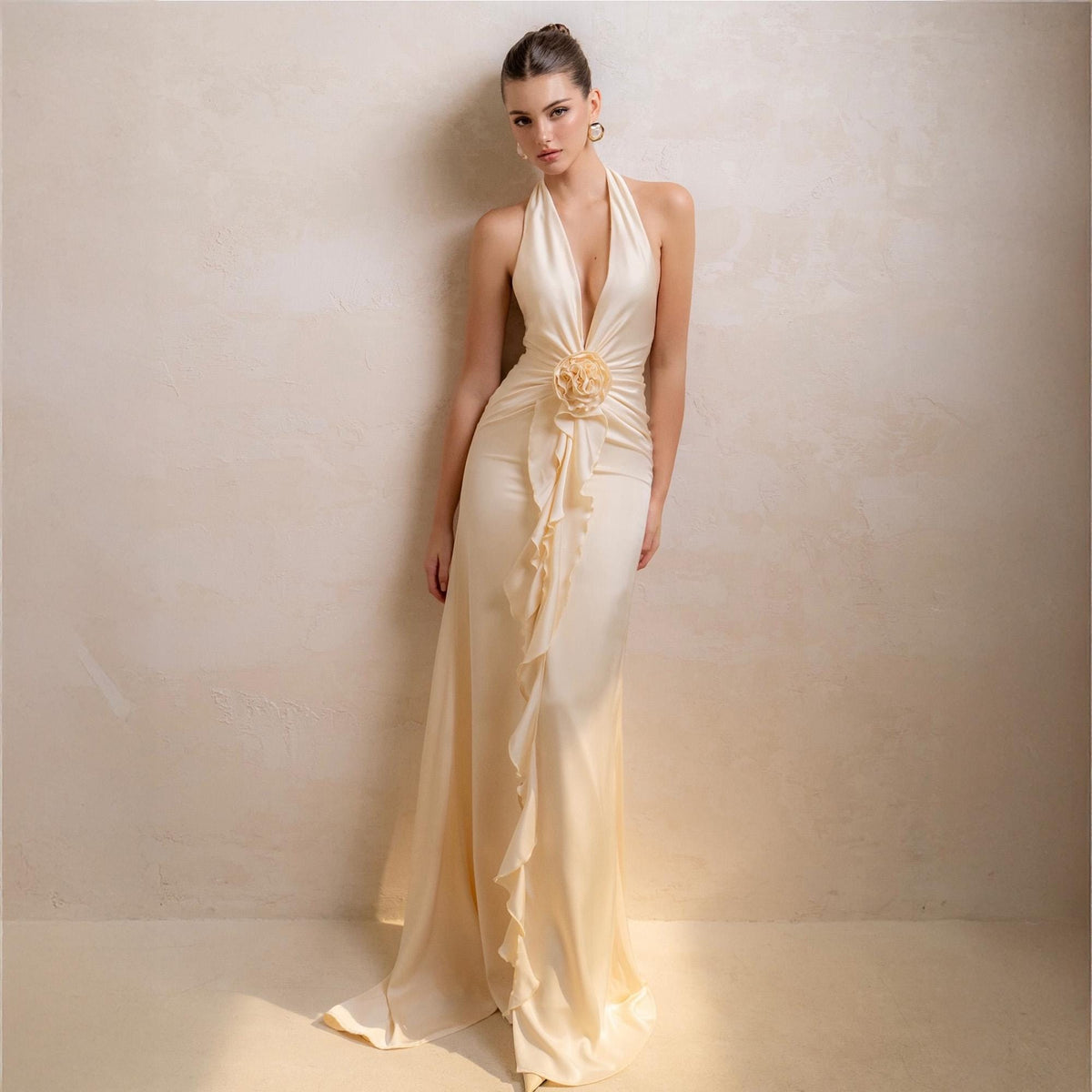 May include: A cream-colored halter neck gown with a plunging neckline and a flowing skirt. The dress features a floral embellishment at the waist and ruffled detailing. The model is standing in front of a neutral-toned wall.