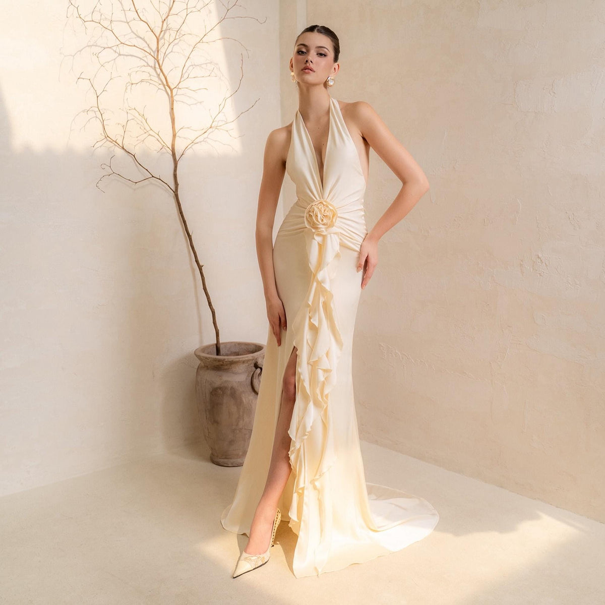 May include: A cream-colored, halter-neck gown with a plunging neckline and a high slit. The dress features a ruffled accent down the front and a floral embellishment at the waist. The model is wearing gold-colored heels.
