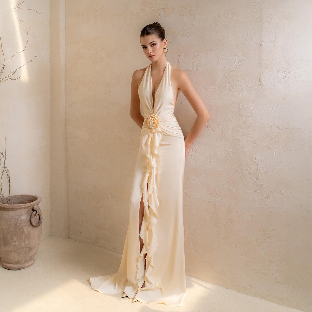 May include: A cream-colored halter neck gown with a plunging neckline and a high slit. The dress features a floral embellishment at the waist and cascading ruffles down the front. The floor-length dress has a flowing silhouette.