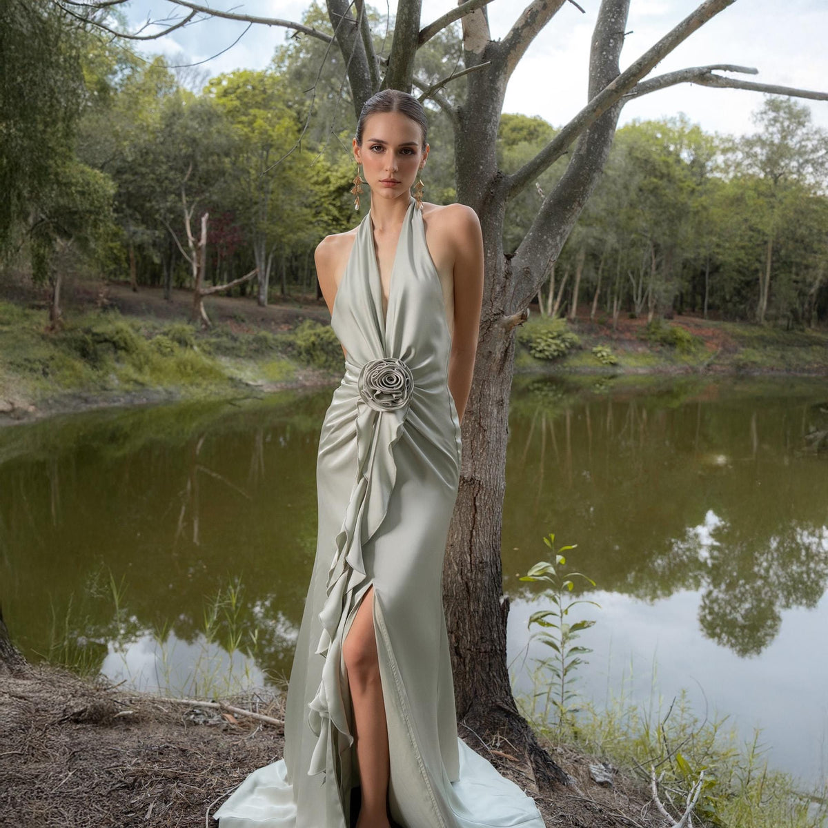May include: A light green, floor-length halter dress with a high slit and a decorative floral embellishment at the waist. The dress features a flowing, ruffled design and is made of a silky material. The background includes a lake and trees.