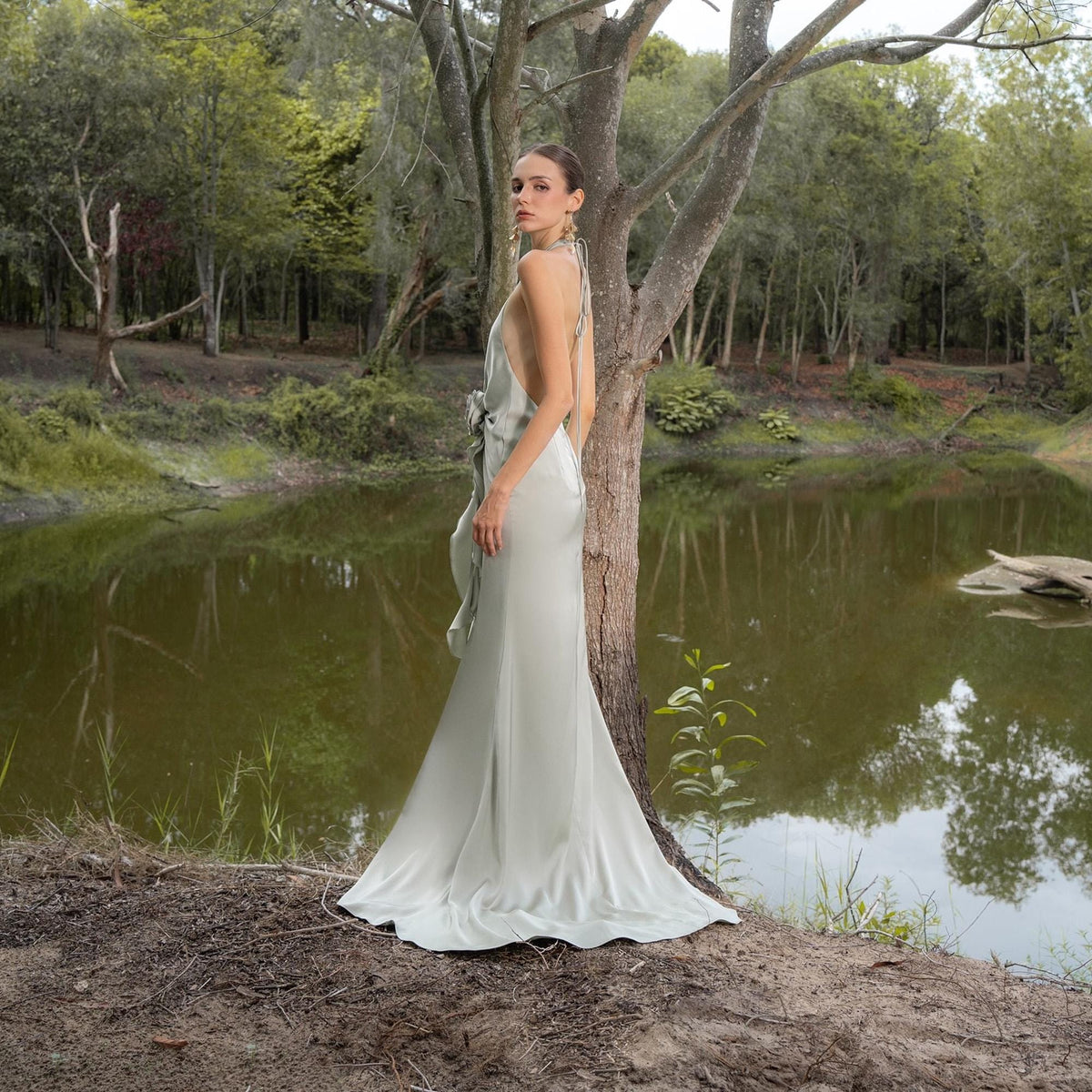 May include: A light green, floor-length gown with a halter neckline and open back. The dress features a bow detail at the waist and a flowing skirt. The model is standing near a tree and a body of water.