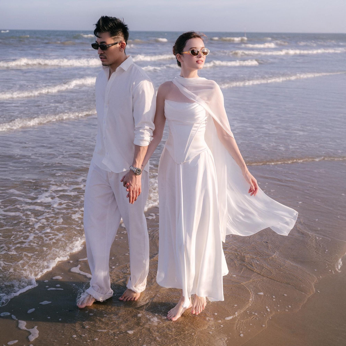 May include: A couple stands on a beach, both dressed in white. The person on the left wears a white button-down shirt and pants, while the person on the right wears a white dress with a flowing cape. Both wear sunglasses.