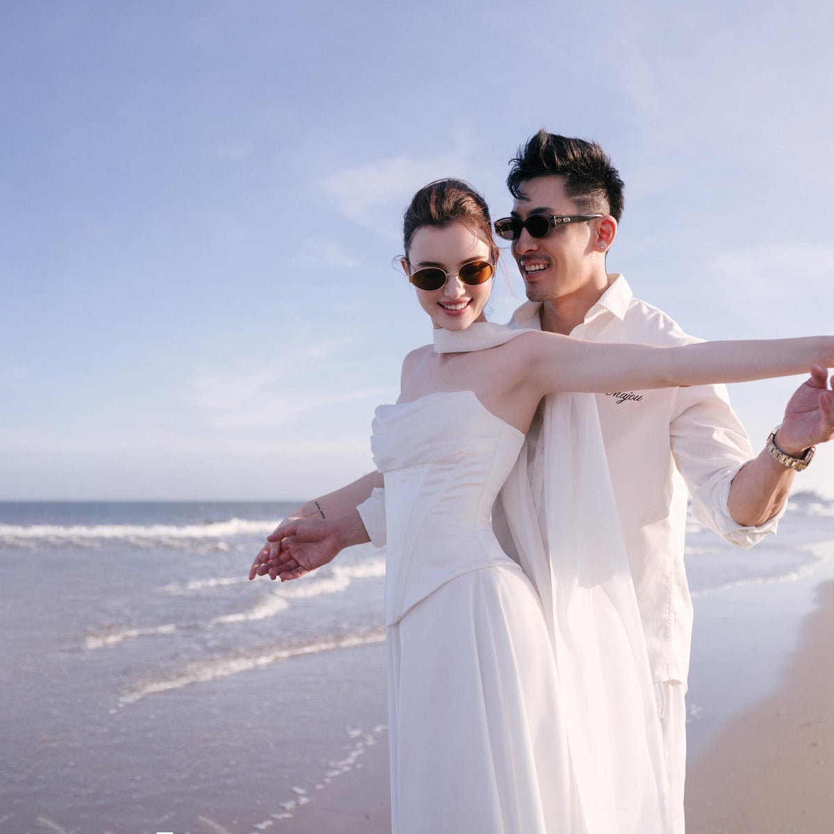 May include: A couple on a beach, the woman in a white strapless dress and the man in a white shirt, both wearing sunglasses. The woman has her arms outstretched. The ocean and sky are in the background.