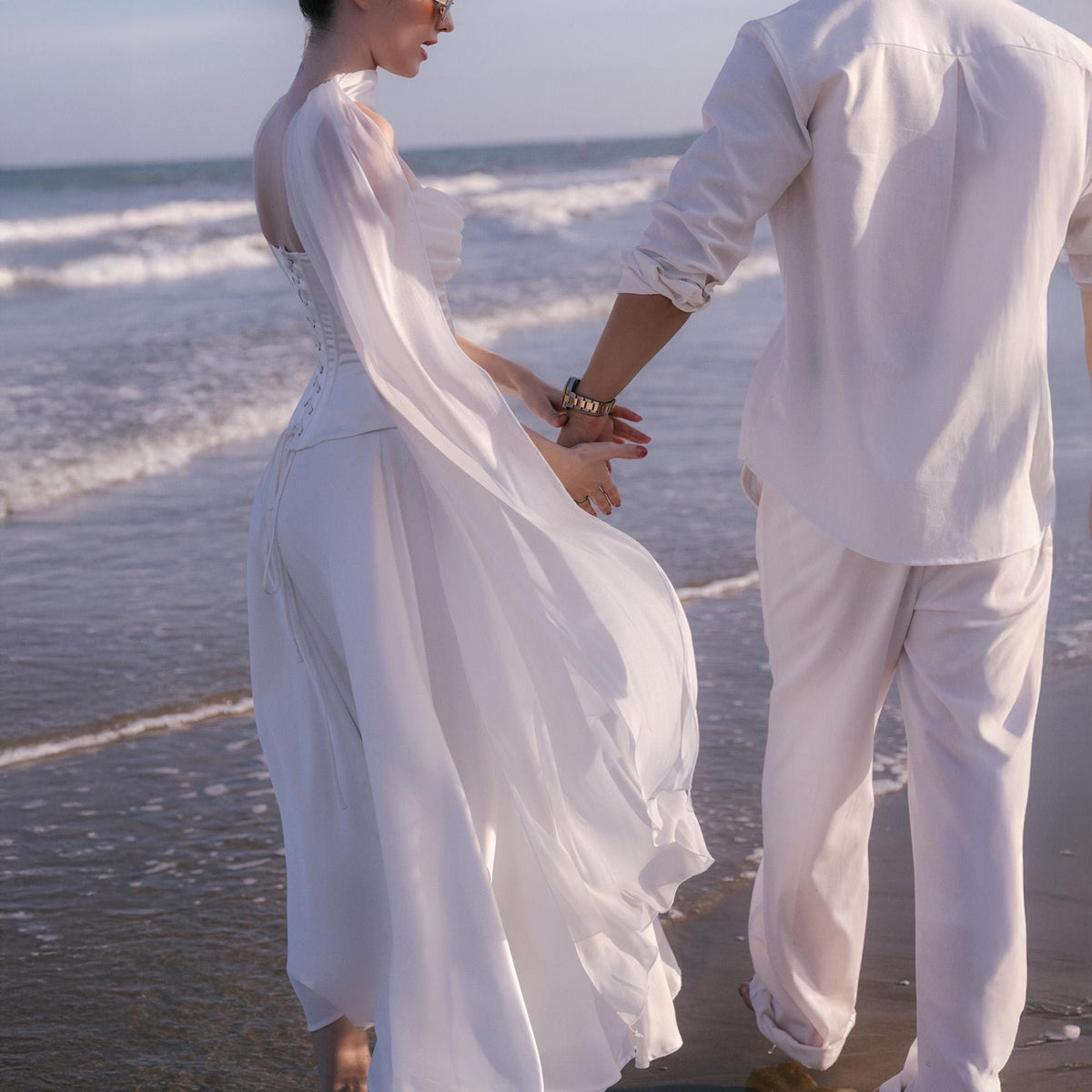 May include: A white wedding dress with a flowing skirt and long sleeves. The dress is paired with a white shirt and pants. The couple is walking on a beach near the ocean.