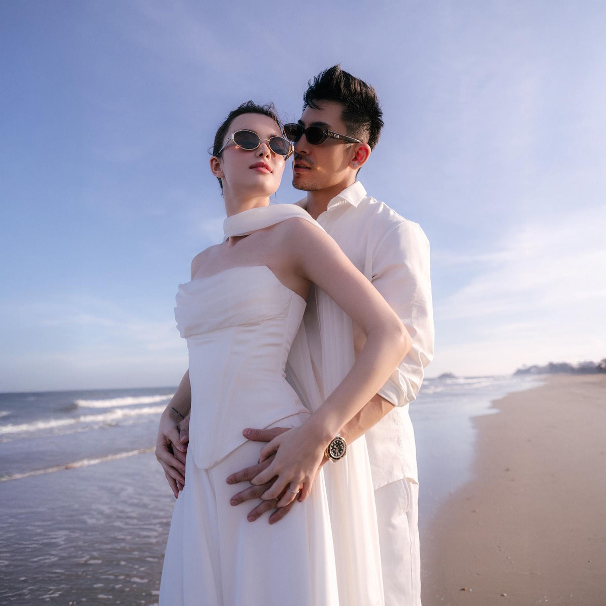 May include: A couple stands on a beach, embracing. The woman wears a white dress and sunglasses, while the man wears a white shirt and sunglasses. The background features the ocean and a clear blue sky. A watch is visible on the man's wrist.