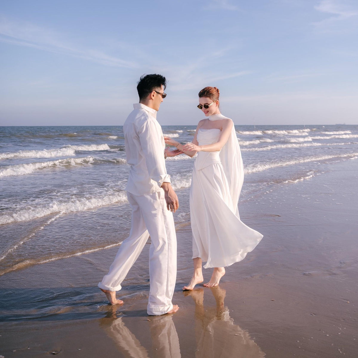May include: A couple in white outfits and sunglasses stand on a beach. The woman wears a white dress with a cape, and the man wears a white shirt and pants. The ocean and sky are visible in the background.