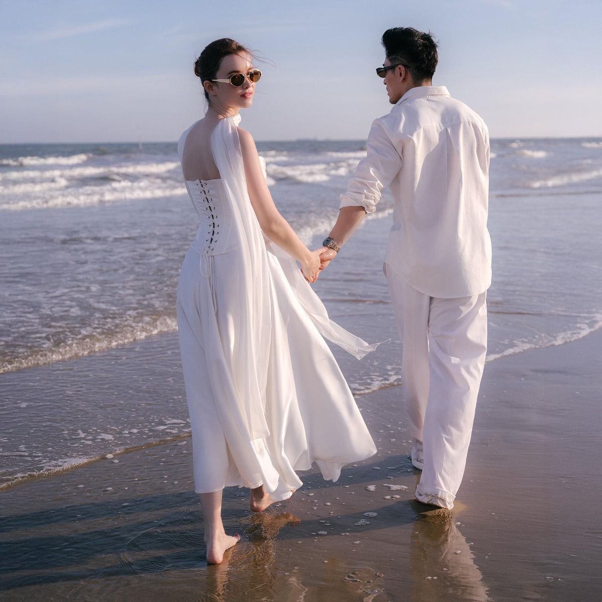 May include: A couple walks along a beach, holding hands. The woman wears a white dress with a corset-style bodice and flowing skirt. The man is in a white shirt and pants. Both wear sunglasses. The scene is bathed in sunlight.