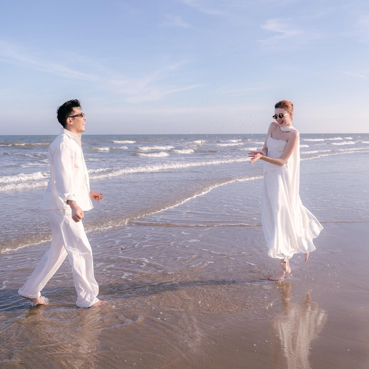 May include: A couple is enjoying a sunny day at the beach. The man is wearing a white button-down shirt and white pants. The woman is wearing a white dress and sunglasses. The ocean and sky are visible in the background.