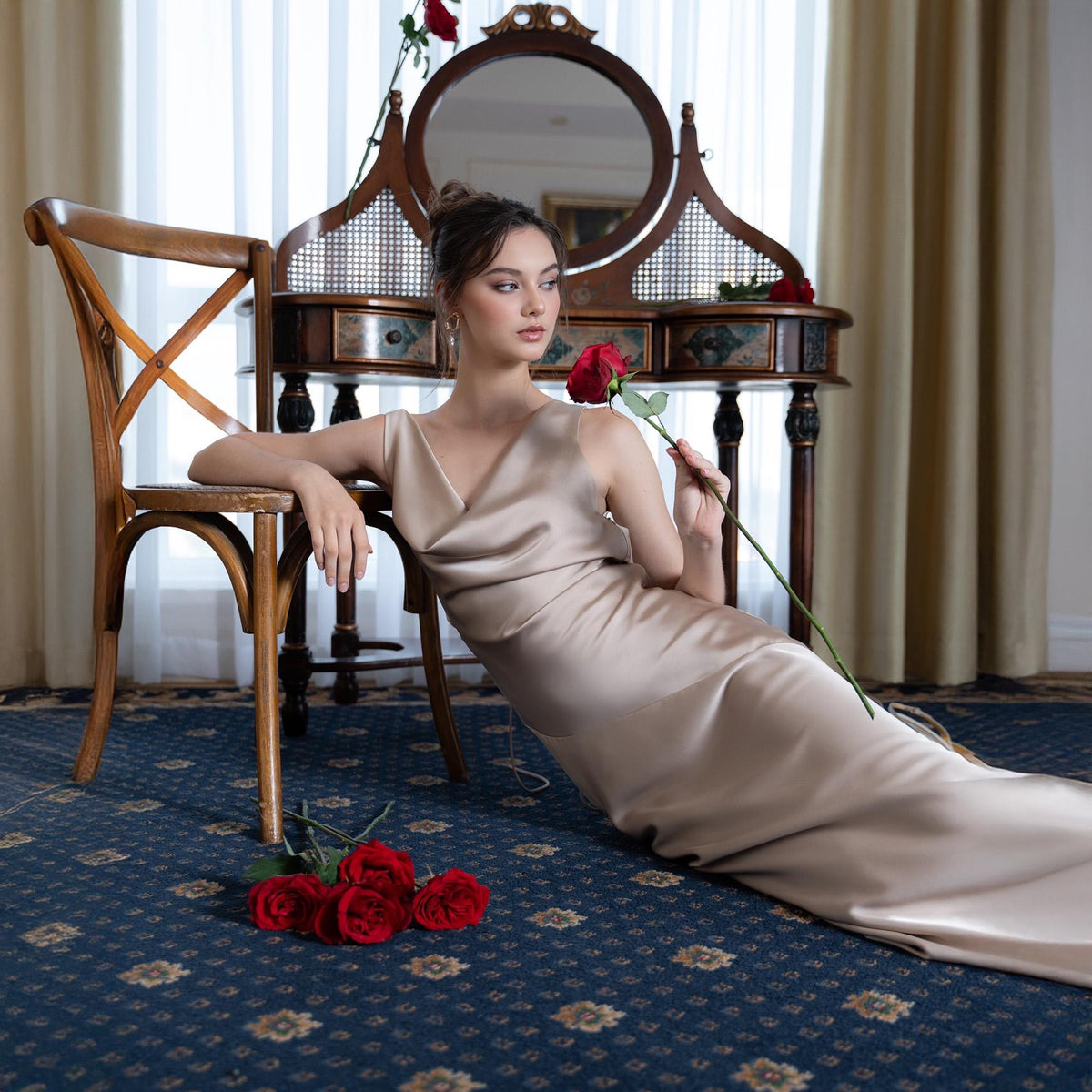 May include: A woman in a champagne-colored satin gown sits near a wooden chair and antique vanity. She holds a red rose, with more roses on the floor. The room has a blue patterned carpet and a window with sheer curtains.