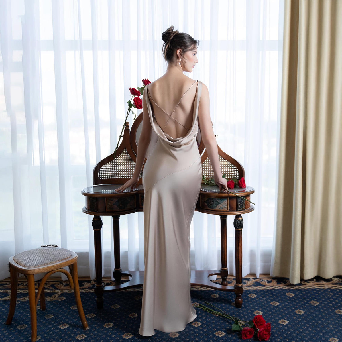 May include: A beige satin gown with a cowl neckline and crisscross back straps. The long dress is worn by a person standing near a vintage vanity table with red roses. The room has a blue patterned carpet and a window with sheer curtains.