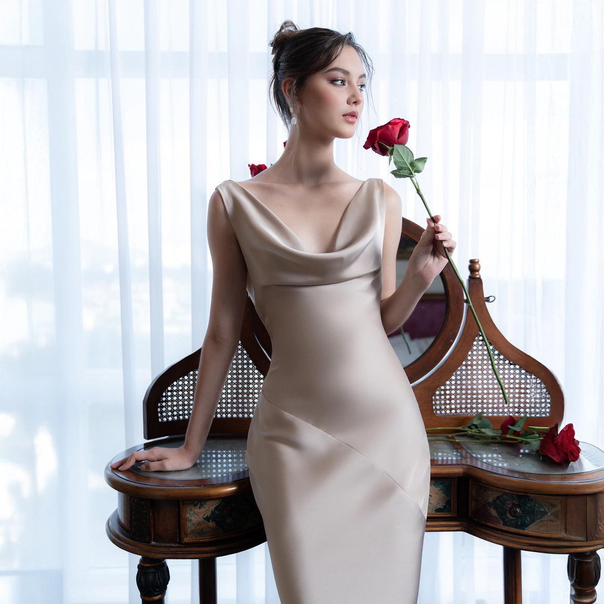 May include: A sleeveless, champagne-colored dress with a cowl neckline. The dress is form-fitting and falls to the floor. A red rose is held in the hand. The background includes a vintage vanity and a sheer white curtain.