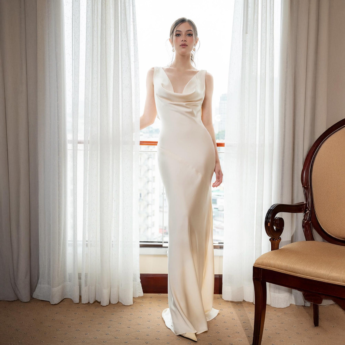 May include: A floor-length, ivory-colored satin gown with a cowl neckline. The sleeveless dress has a form-fitting silhouette that gently flares out towards the hem. The model is standing near a window with sheer white curtains.