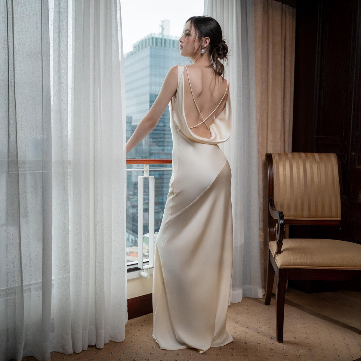 May include: A cream-colored, floor-length gown with a cowl back and spaghetti straps. The dress has a flowing silhouette and is made of a silky material. The model is standing near a window with white curtains, looking out at a cityscape.