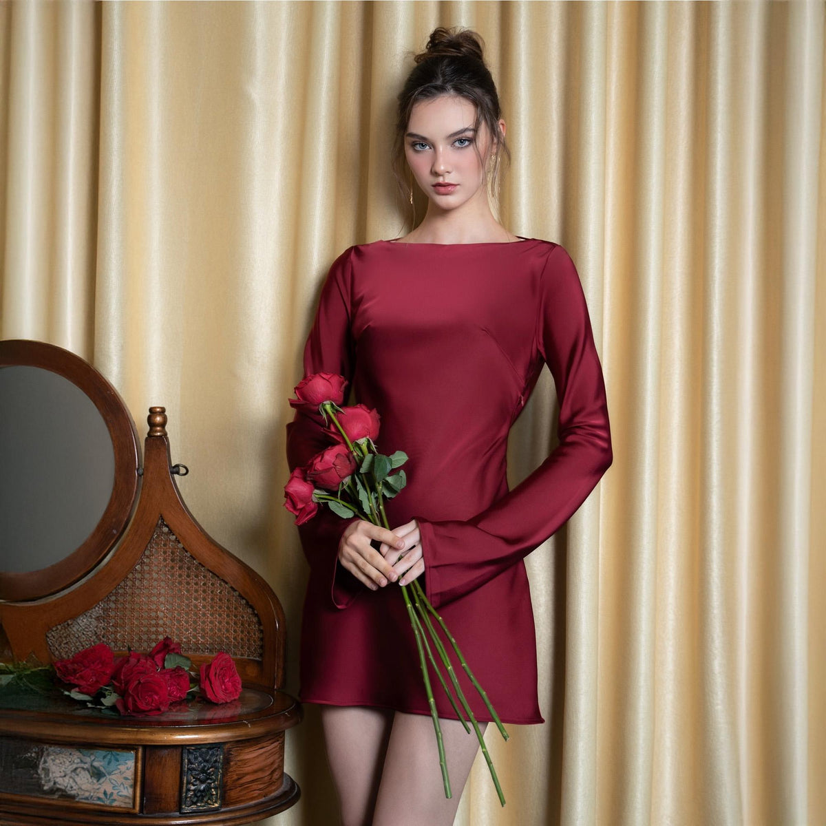 May include: A burgundy-colored, long-sleeved dress with a boat neckline. The dress is made of a shiny fabric and is held by a person. The person is holding a bouquet of red roses. A vintage vanity mirror is in the background.