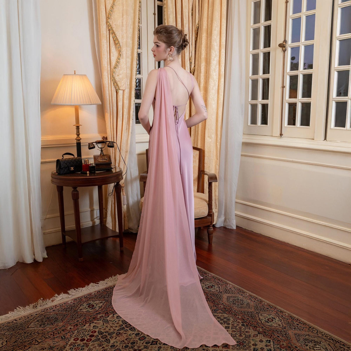 May include: A light pink formal gown with a flowing cape detail. The dress features a draped neckline and a low back with delicate strap detailing. The model is standing in a room with a vintage aesthetic, with a lamp and a small table in the background.