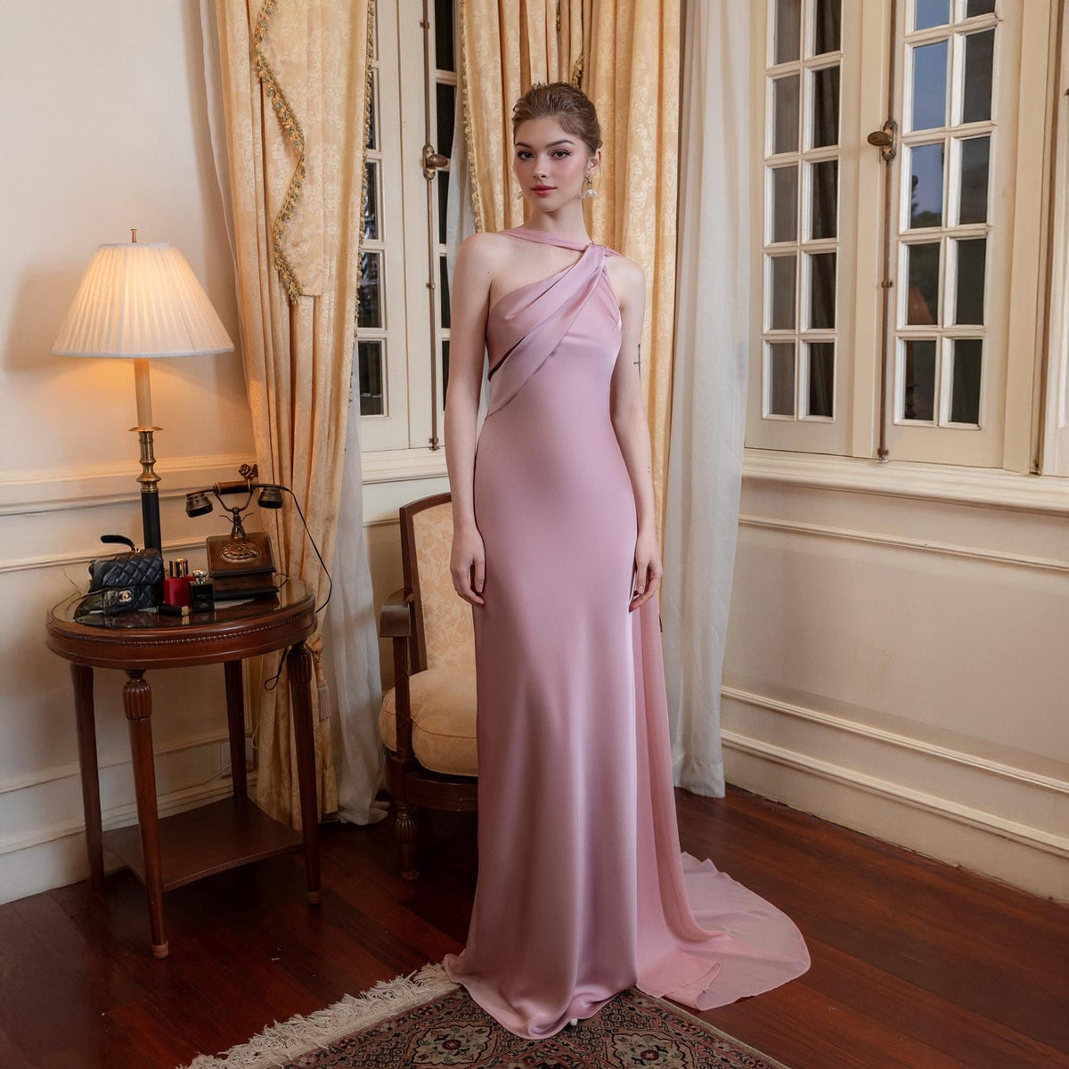 May include: A dusty rose, one-shoulder formal gown with a flowing skirt and a draped bodice. The dress has a subtle sheen and a long, elegant train. The model is standing in a room with a vintage aesthetic, with a table and a chair.