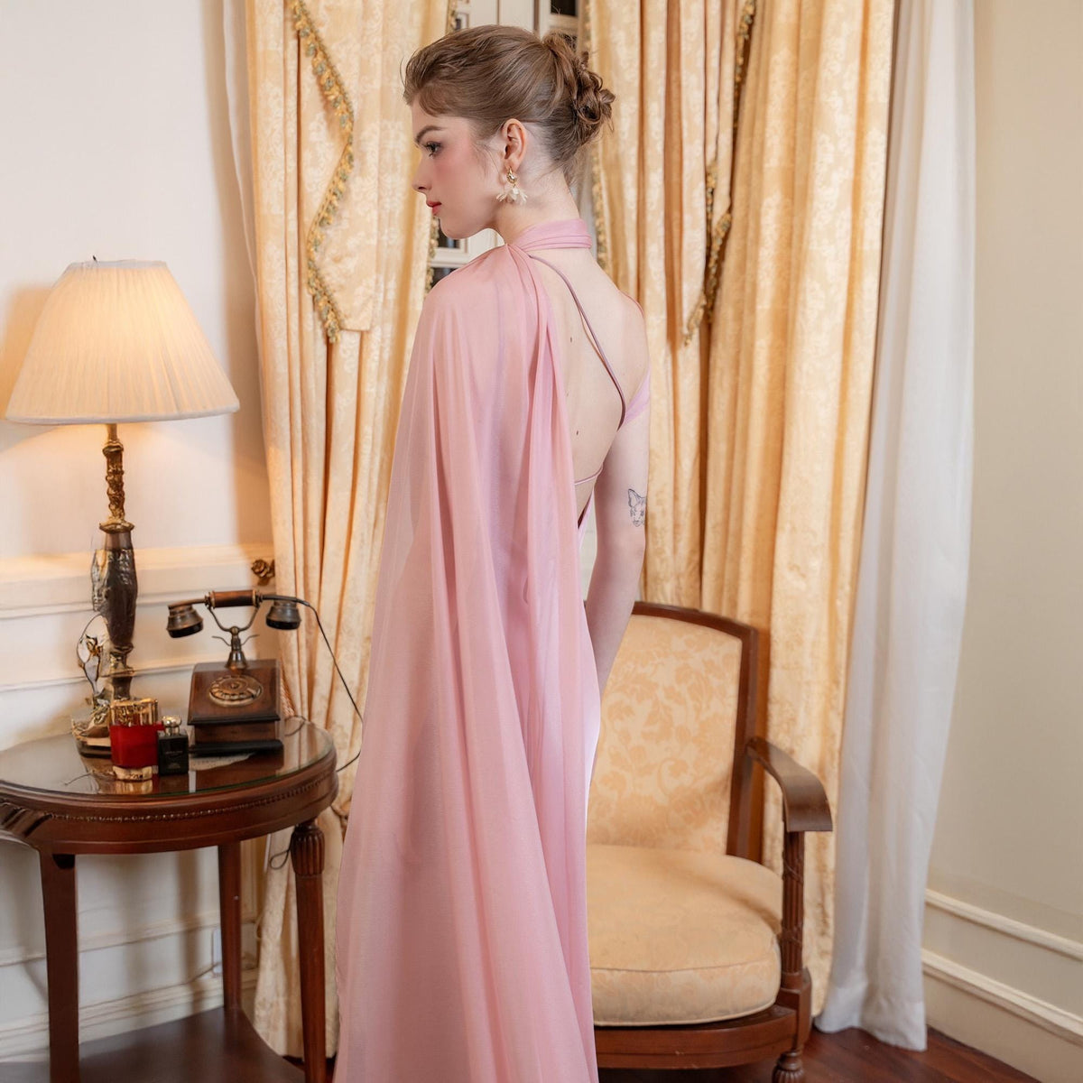 May include: A light pink, flowing evening gown with a draped cape detail. The dress features a halter neckline and a low back. The model is standing in front of a vintage lamp and a wooden side table.