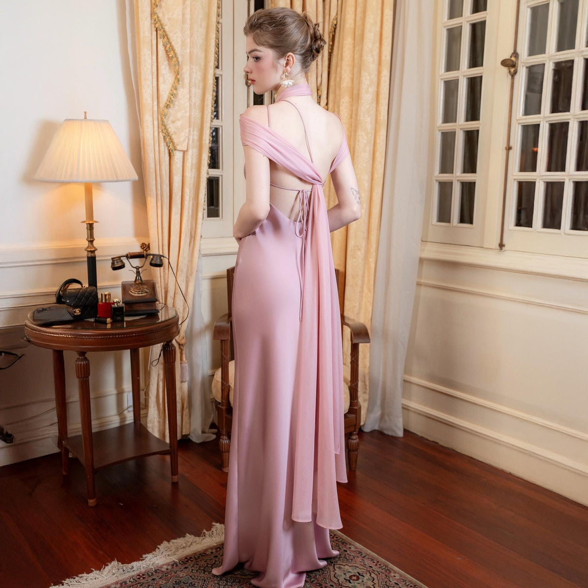 May include: A floor-length, pale pink formal gown with a halter neckline and open back. The dress features draped fabric over the shoulders and a flowing train. The gown is made of a silky material and is suitable for special occasions.