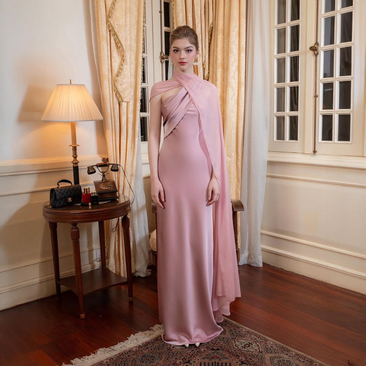 May include: A floor-length, pale pink satin gown with a draped cape. The dress features a halter neckline and a flowing skirt. The model is standing in a room with a vintage aesthetic, with a lamp and a small table.