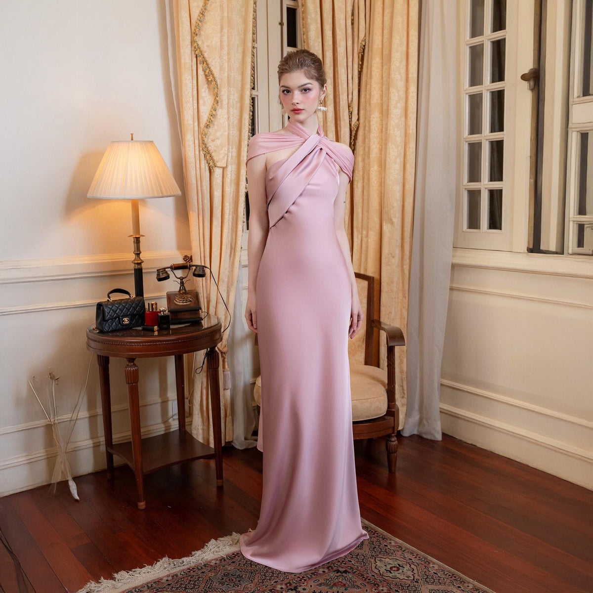 May include: A floor-length, pale pink gown with a halter neckline and a fitted silhouette. The dress features a draped bodice and a subtle train. The background includes a vintage-style room with a lamp, table, and curtains.