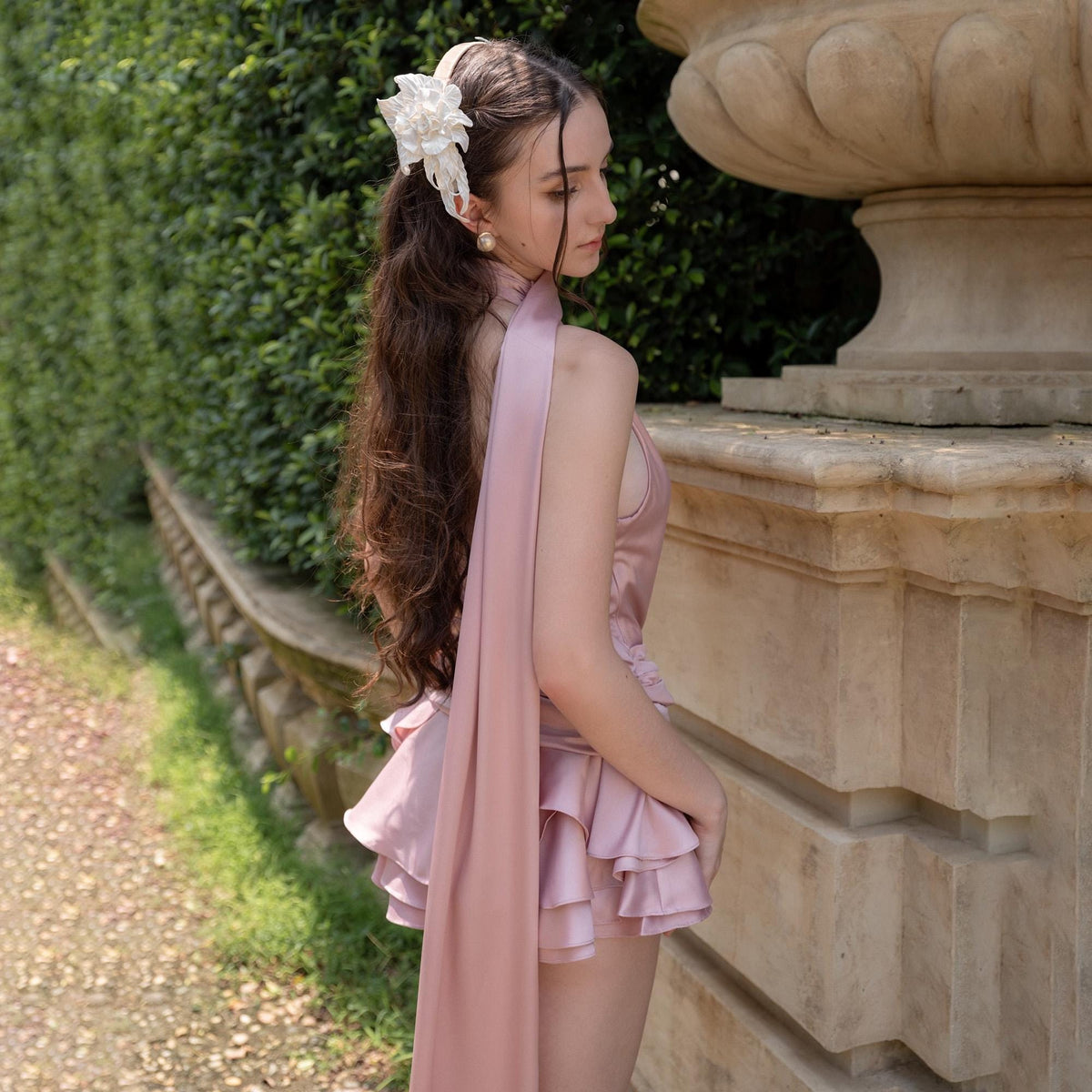 May include: A light pink satin romper with layered ruffles and a halter neckline. A long, flowing sash drapes from the back. The model is wearing a white floral hair accessory and pearl earrings. The background features a stone wall and greenery.