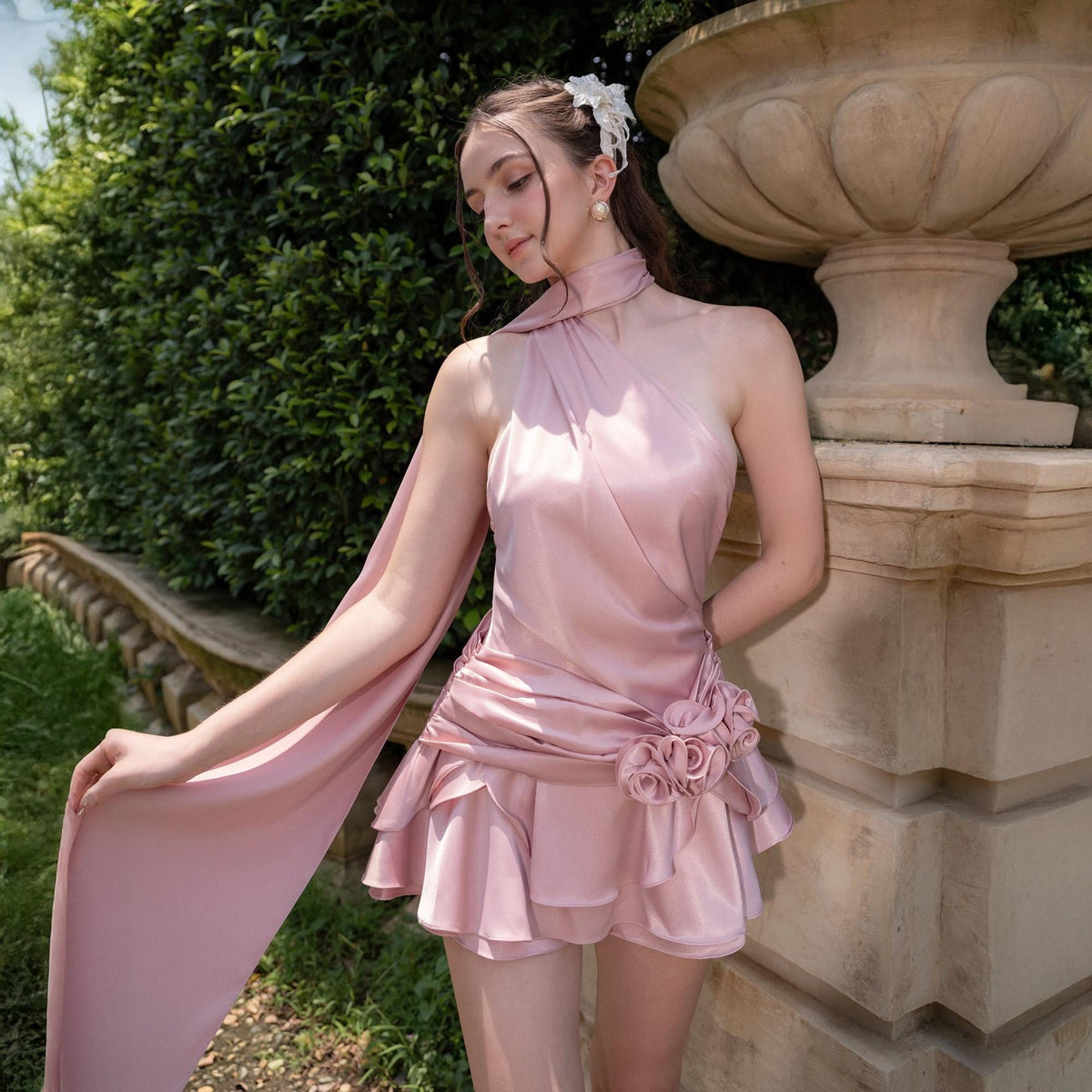 May include: A light pink satin dress with a halter neckline and a flowing, draped design. The dress features a layered skirt and a floral embellishment at the waist. The model is wearing a white hair accessory and pearl earrings.