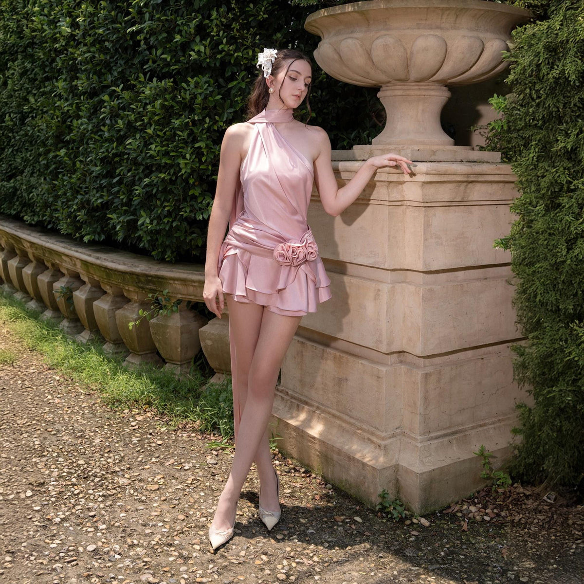 May include: A light pink satin dress with a halter neckline and a ruffled skirt. The dress features a floral embellishment at the waist. The model is wearing white heels and a white flower in their hair. The background includes a stone wall and greenery.