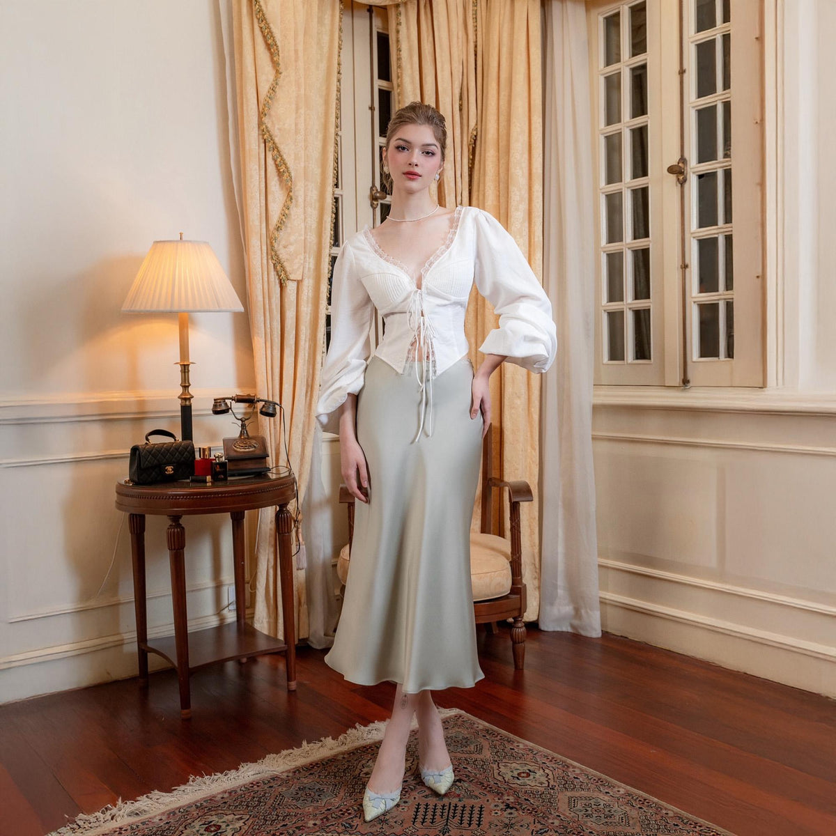 May include: A woman wearing a white blouse with lace trim and a light green satin skirt. The blouse has long, puffy sleeves and a tie-up front. The skirt is a midi length. The woman is standing in a room with a vintage aesthetic.