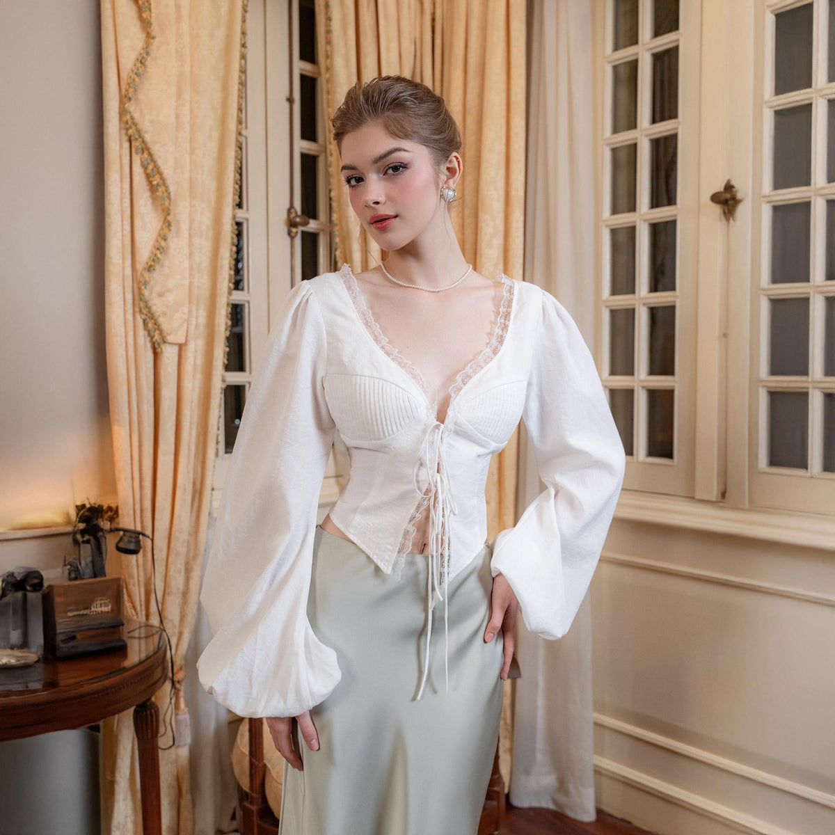May include: A white blouse with long, voluminous sleeves and a lace-trimmed, plunging neckline. The bodice features vertical pleats and a tie-up front. The blouse is paired with a light green satin skirt, creating a vintage-inspired ensemble.