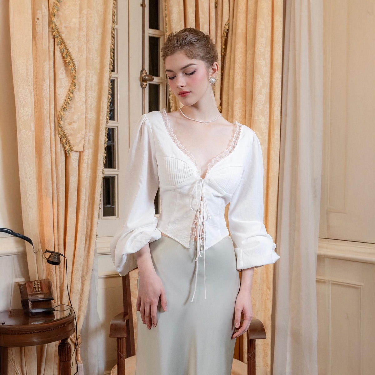 May include: A white blouse with long sleeves and a lace-trimmed, V-neckline. The blouse features a corset-style bodice with a tie-up front. The model is wearing a light green satin skirt. The background includes a window and curtains.