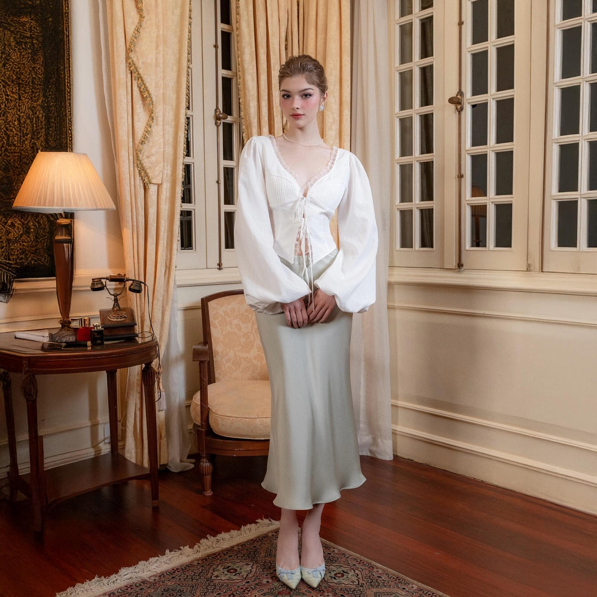 May include: A woman's outfit featuring a white, long-sleeved blouse with a lace-trimmed neckline and a light green satin skirt. The blouse has a tie-up front and the skirt falls to the ankles. The outfit is paired with matching heels.
