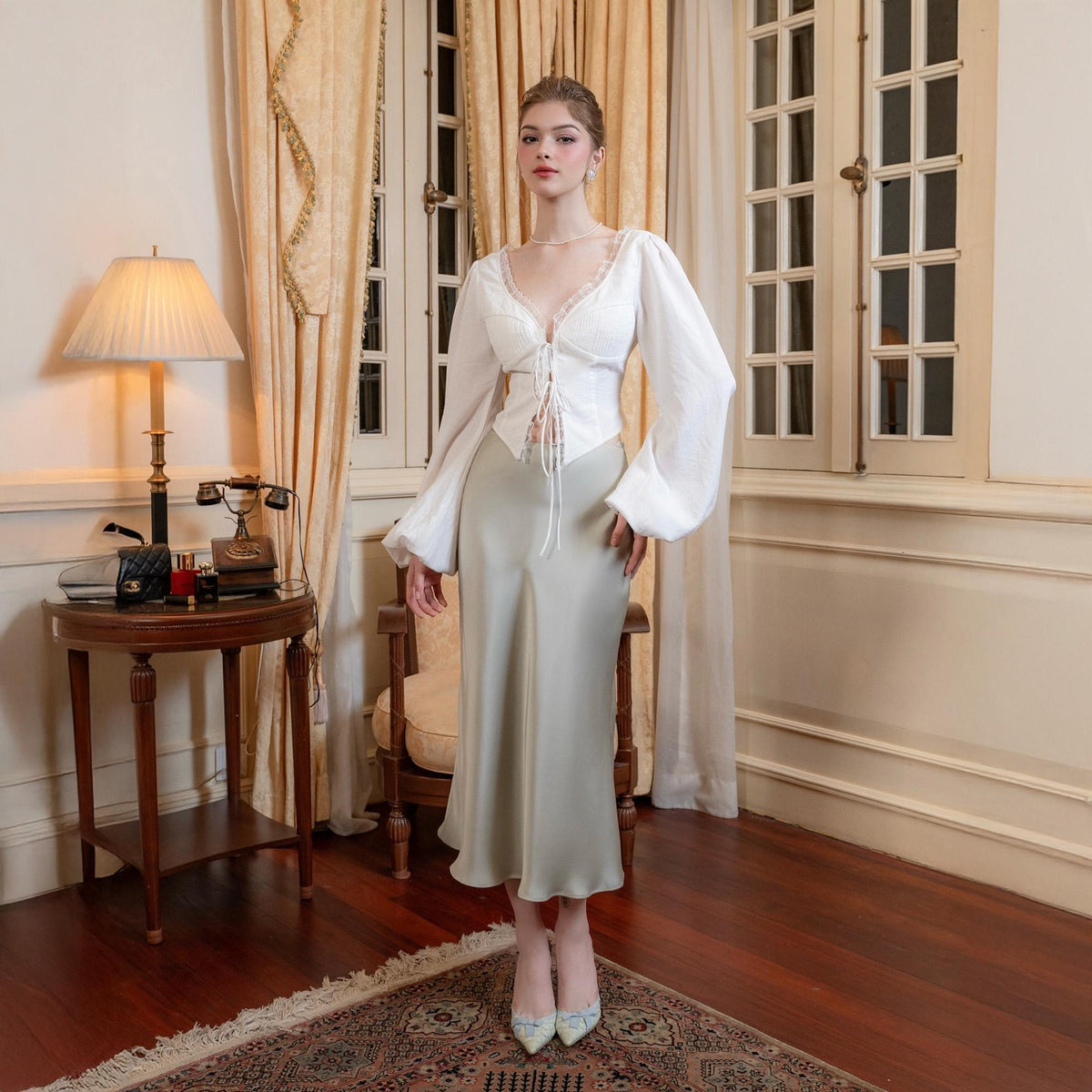 May include: A woman wearing a white long-sleeve blouse with lace trim and a light green satin midi skirt. The blouse has a tie-up front and the skirt falls to mid-calf. She is wearing matching heels.