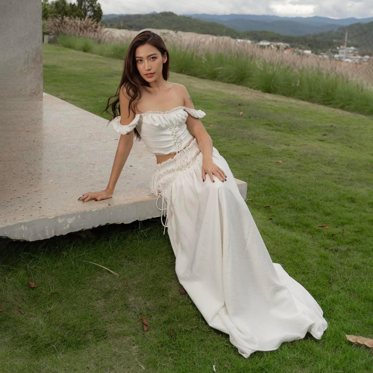May include: A white two-piece outfit featuring an off-the-shoulder crop top and a high-waisted maxi skirt. The top has ruffled sleeves and a drawstring detail. The skirt has a textured pattern and a flowing silhouette. The outfit is worn outdoors.
