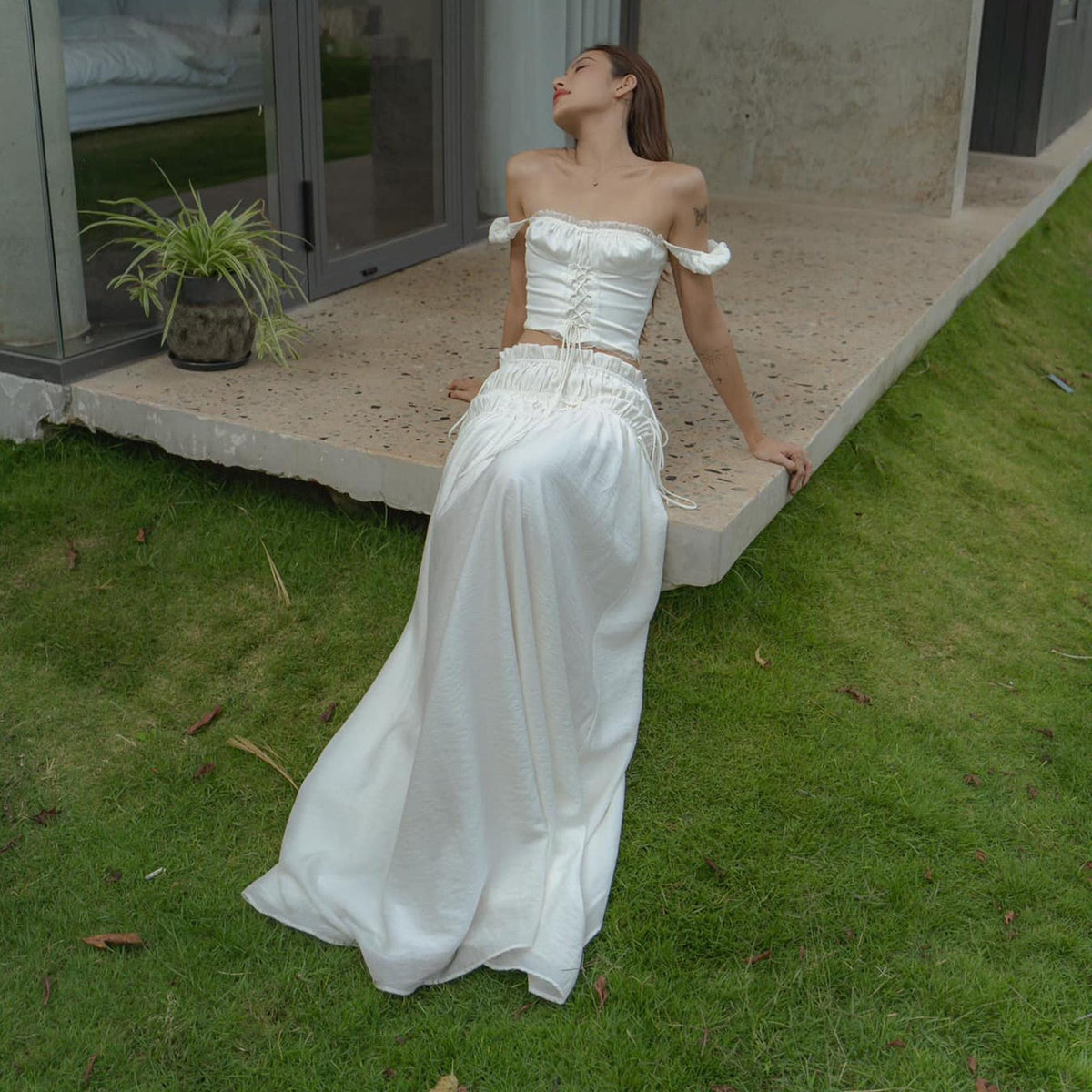May include: A white satin two-piece outfit. The top is a corset style with off-the-shoulder sleeves and a lace trim. The skirt is a long, flowing design. The outfit is worn outdoors on a concrete surface with green grass.