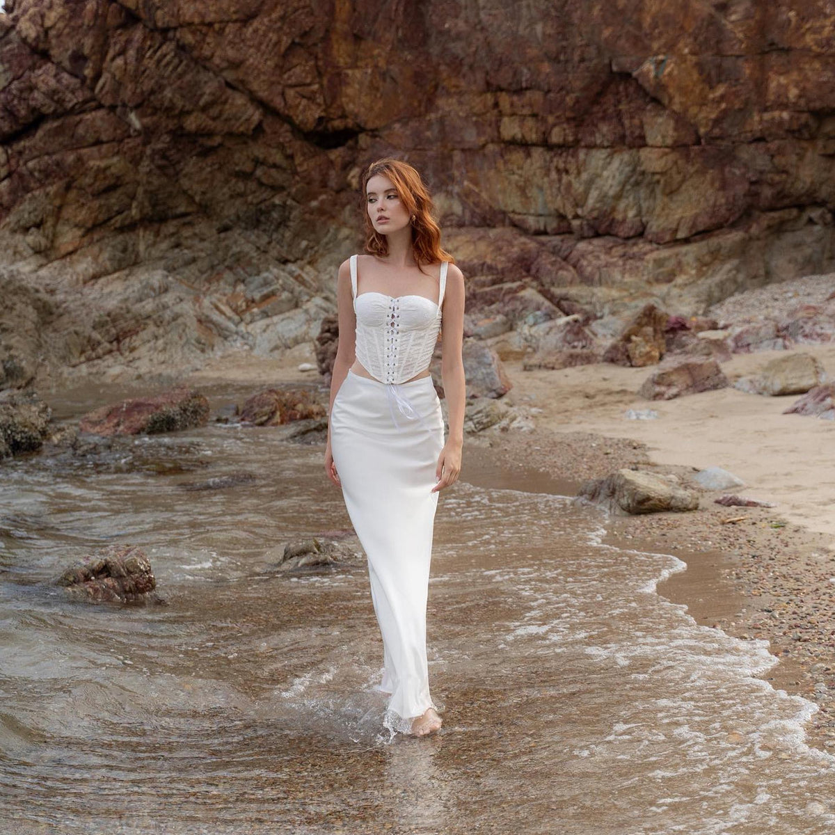 May include: A white corset top with spaghetti straps and a matching white skirt. The corset features a lace-up front and is paired with a long, flowing skirt. The outfit is worn on a beach with a rocky background.