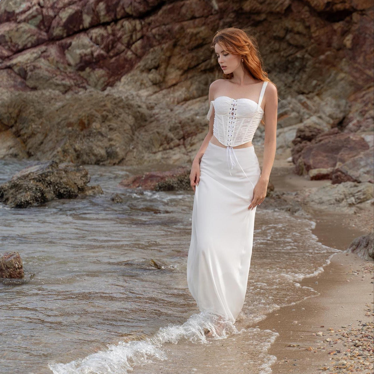 May include: A white corset top with spaghetti straps and a matching white skirt. The outfit is worn on a beach with the ocean in the background. The corset has a lace-up front.