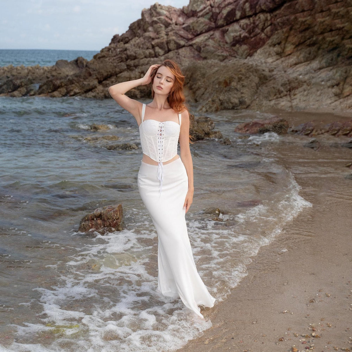May include: A white corset top with spaghetti straps and a lace-up front, paired with a matching white, floor-length skirt. The outfit is worn on a beach with the ocean in the background. The corset has a structured bodice.