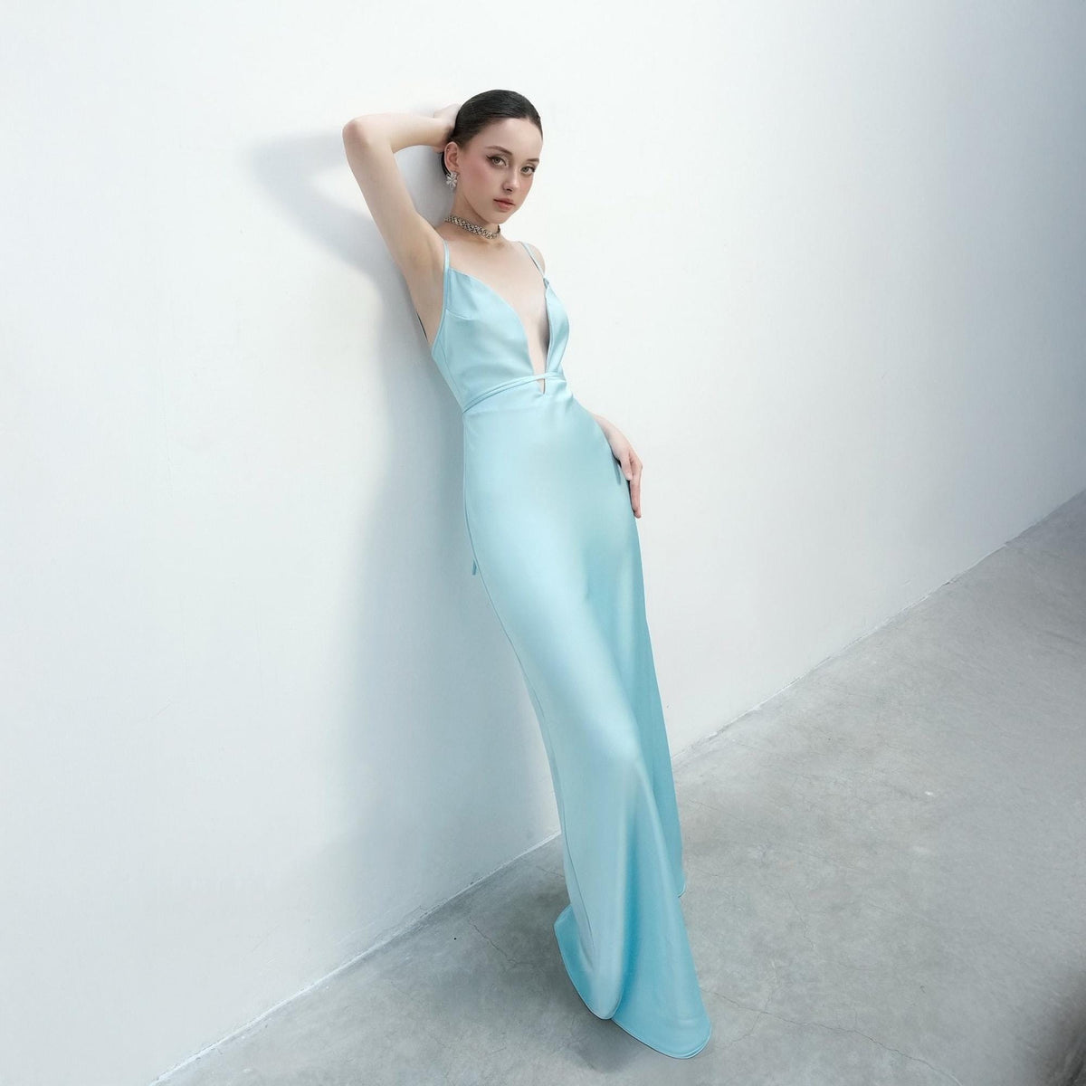 May include: A light blue satin gown with a plunging neckline and spaghetti straps. The dress has a fitted bodice and a flowing skirt. The model is leaning against a white wall, with one arm raised and the other at their side.