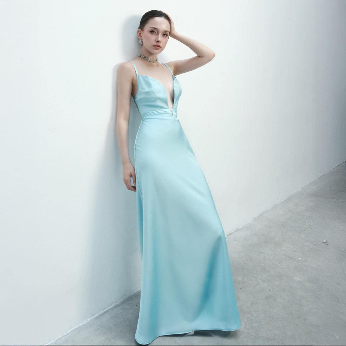May include: A light blue, floor-length satin gown with a plunging neckline and spaghetti straps. The dress has a fitted bodice and a flowing skirt. The model is leaning against a white wall.
