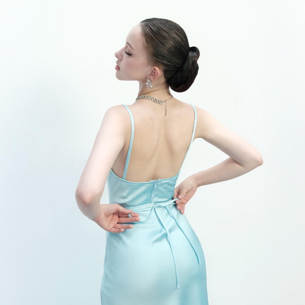 May include: A light blue satin slip dress with spaghetti straps and a low back. The dress features a tie at the waist and a subtle sheen. The model is wearing a silver necklace and earrings, with their hair up in a bun.