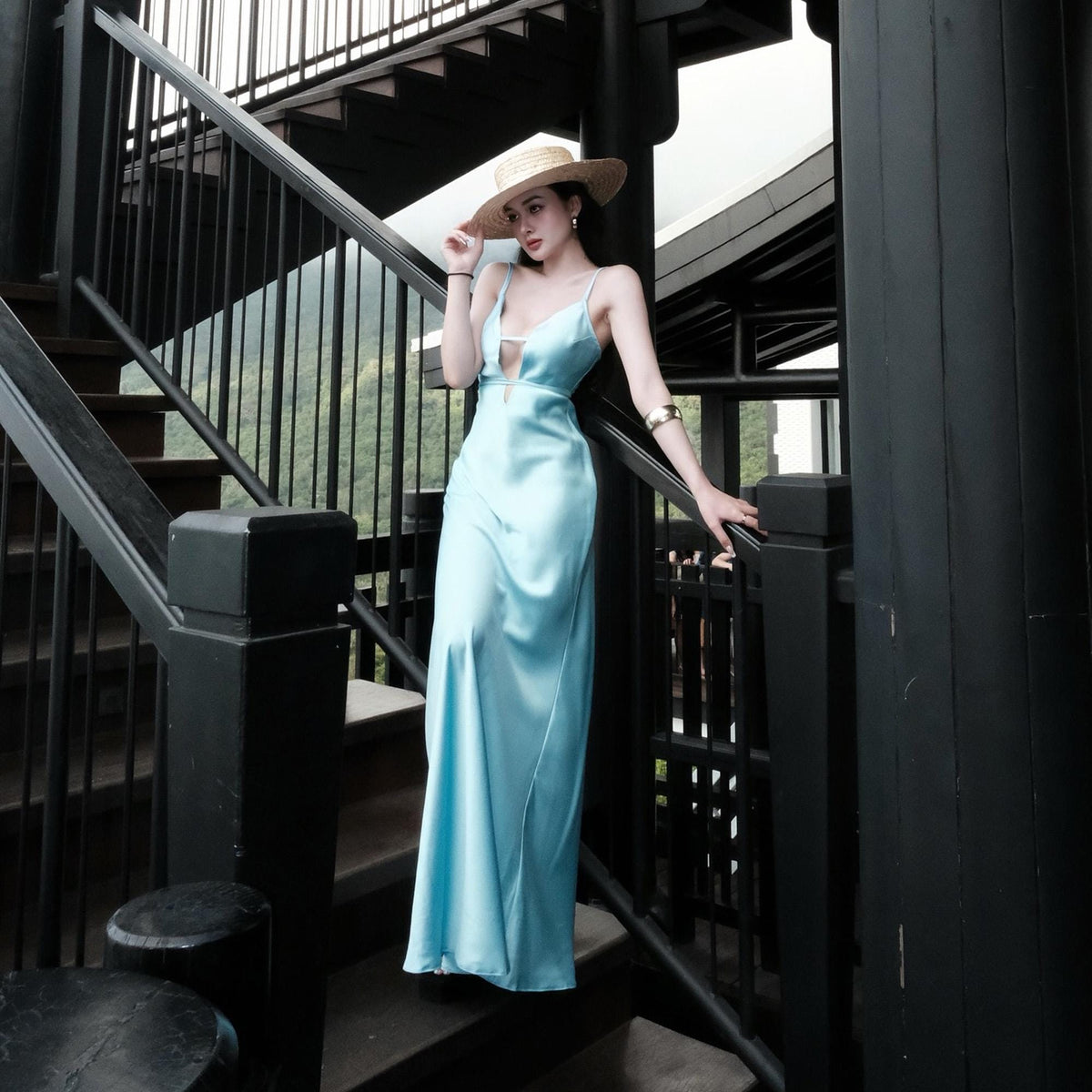 May include: A light blue satin maxi dress with a plunging neckline and spaghetti straps. The dress is worn with a straw hat and gold bracelet. The person is standing on a black staircase with a black railing.