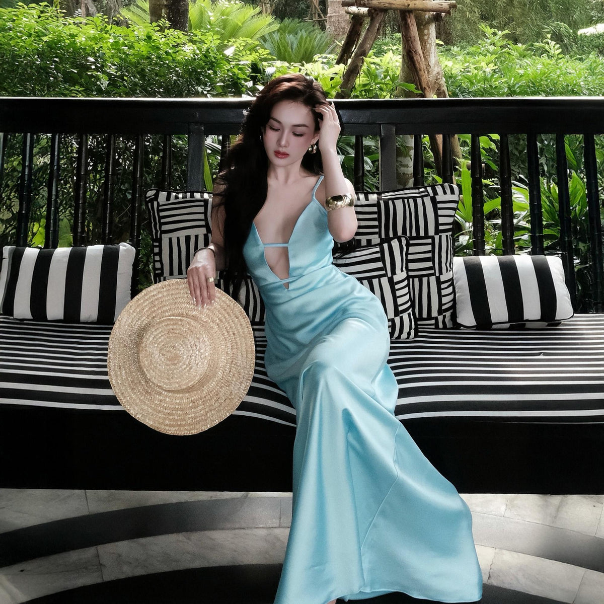 May include: A light blue satin maxi dress with a plunging neckline and spaghetti straps. A straw sun hat is held in the hand. The background features a black and white striped bench with decorative pillows and lush greenery.