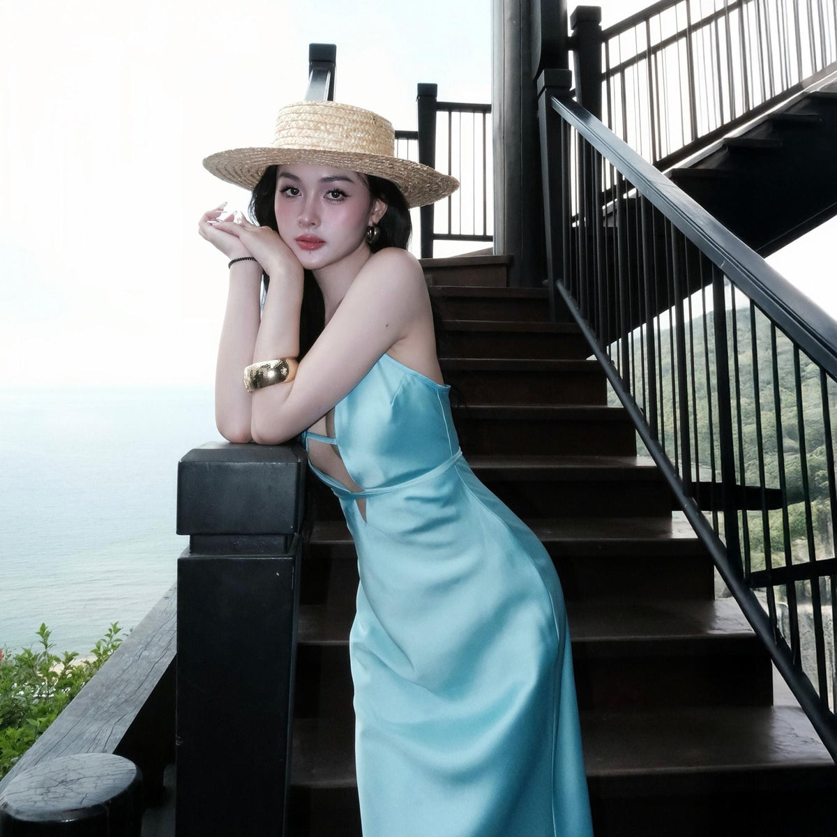 May include: A light blue satin dress with spaghetti straps and a keyhole neckline. The person is wearing a straw hat with a wide brim and a gold bracelet. The background includes a wooden staircase and a view of the ocean.