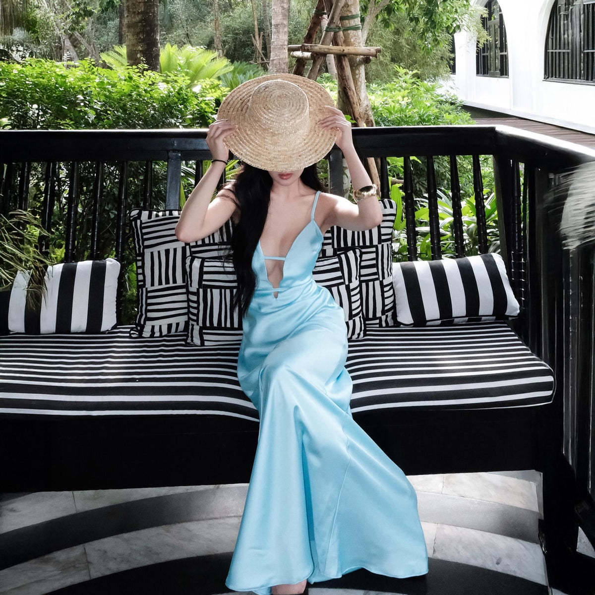 May include: A light blue satin maxi dress with a plunging neckline. The dress is worn with a straw sun hat. The background includes a black and white striped bench with decorative pillows and lush greenery.
