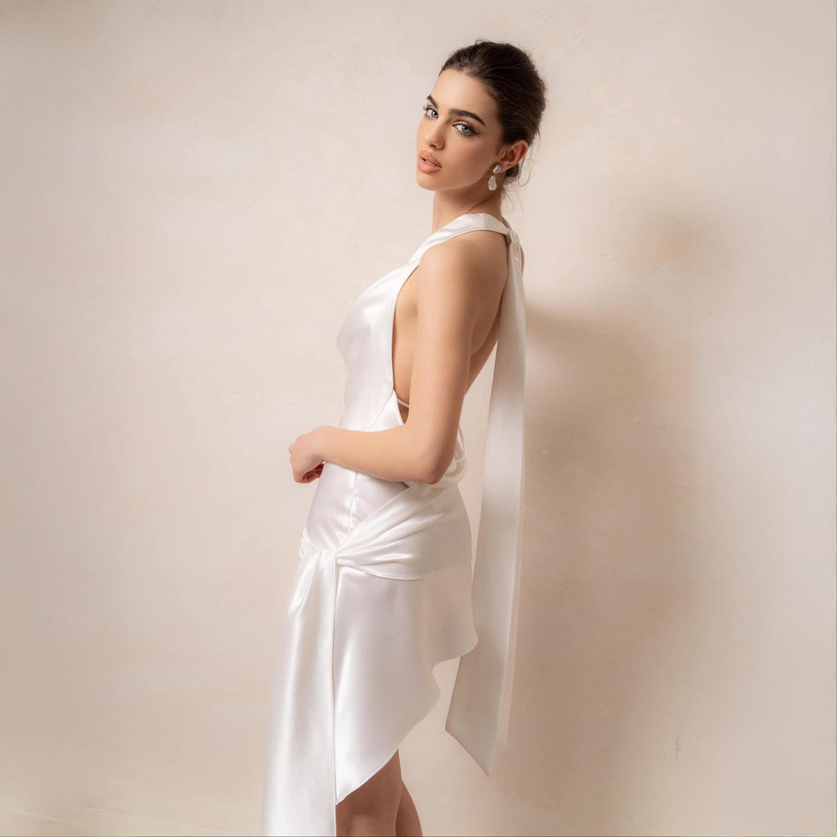 May include: A white satin dress with a draped neckline and a flowing, asymmetrical skirt. The dress features a knotted detail at the waist and a long, flowing sash that drapes down the back. The dress is a formal wear item.