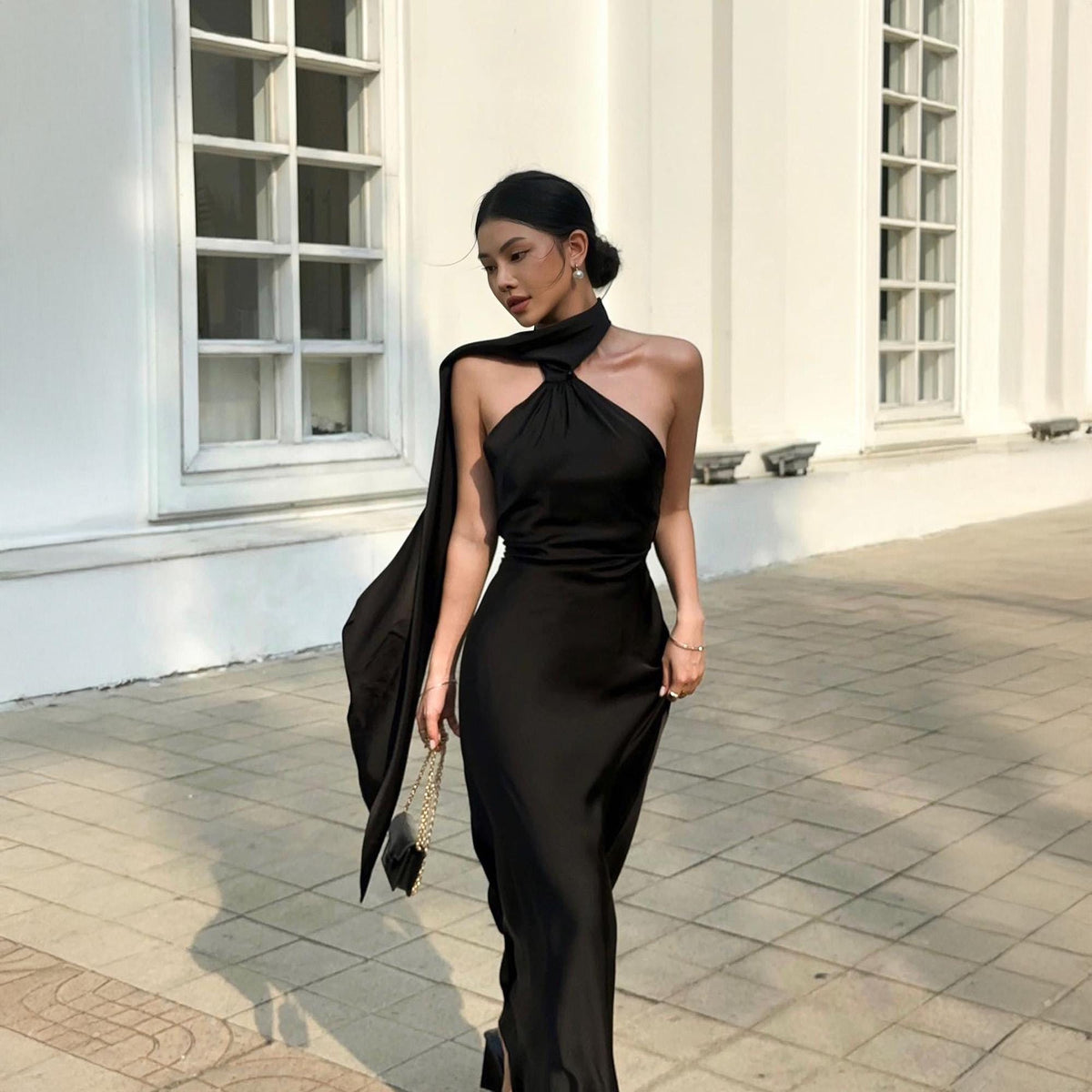 May include: A black, floor-length halter dress with a flowing scarf detail. The dress features a high neckline and a fitted silhouette. A small black purse with a gold chain is held in the hand. The background is a white building with large windows.