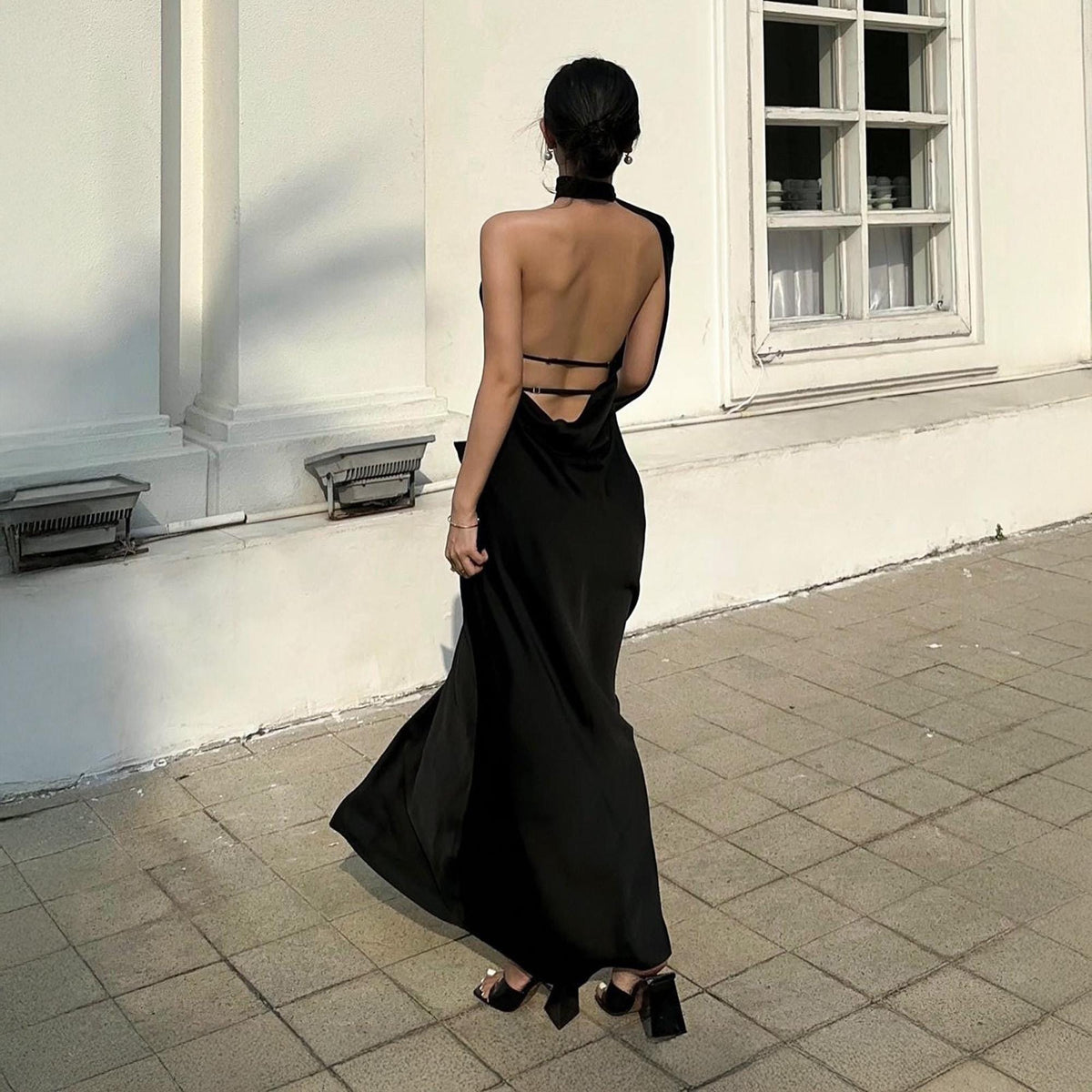 May include: A black, backless maxi dress with a halter neckline and a flowing skirt. The dress is paired with black heeled sandals. The image is taken outdoors, with a white building in the background.