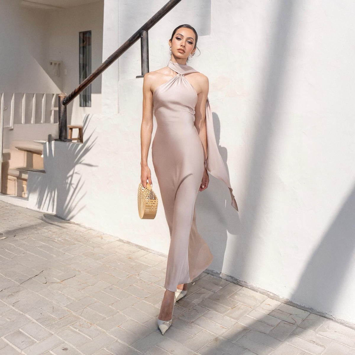 May include: A light pink, floor-length dress with a halter neckline and a flowing scarf detail. The dress is paired with white heels and a woven handbag. The model is standing on a brick walkway next to a white wall.