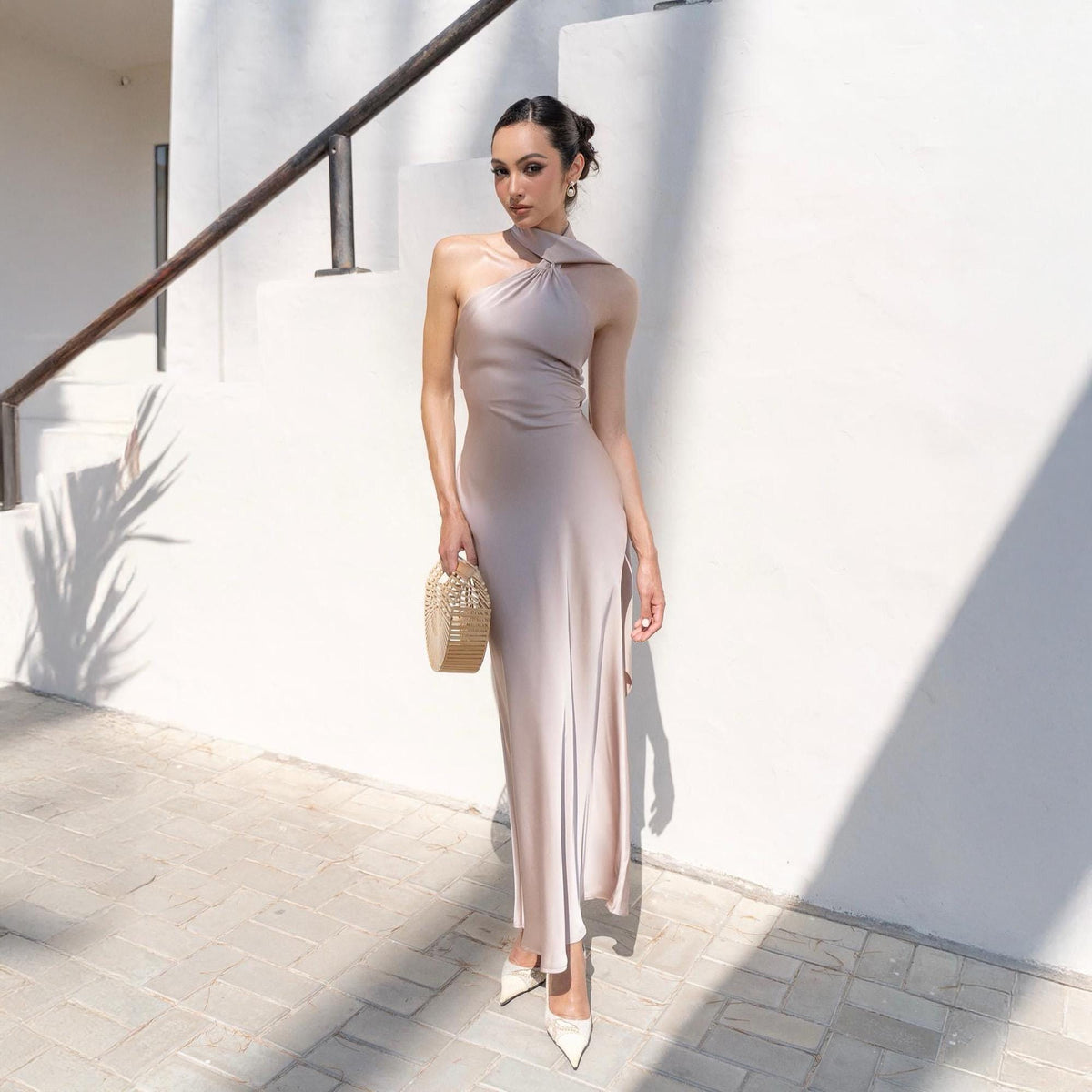 May include: A light beige, floor-length dress with a halter neckline and a side slit. The dress is paired with white pointed-toe heels and a small, woven gold handbag. The background is a white wall with shadows.