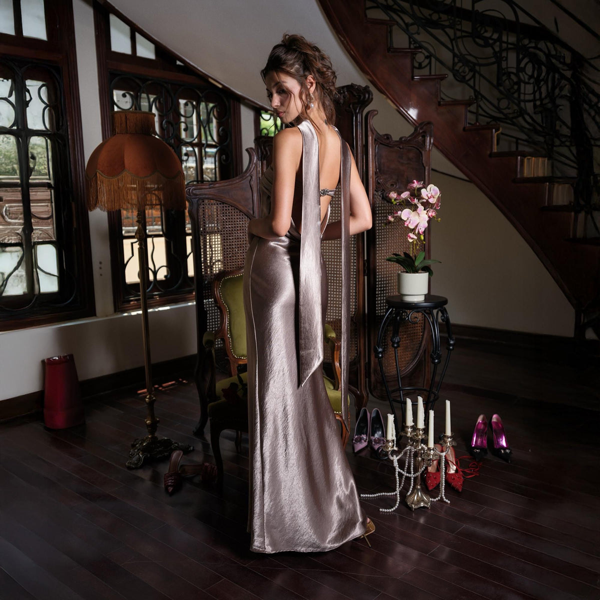 May include: A long, shimmering, taupe-colored gown with a flowing design. The dress features a halter-style neckline and long, draping fabric that extends down the back. The image is set in a room with a vintage aesthetic, with a decorative screen and a candelabra.